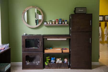 a large play itchen made of wood. A round mirror is on the wall to one side. A small basket of neatly arranged stuffed animals is underneath.