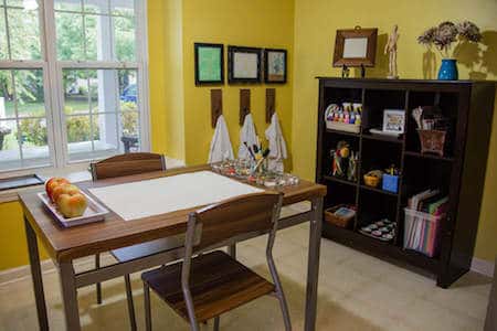 a table with art supplies and apples on top. An aterlier full of other supplies is in the background. Three paint shirts hang on pegs next to the atelier.